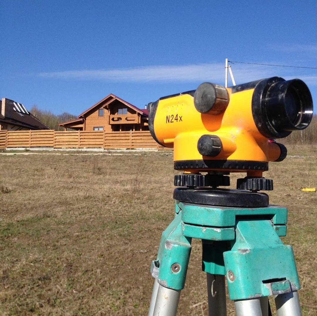 Żółty niwelator optyczny N24x na zielonym statywie, ustawiony na trawiastym terenie z widokiem na drewniany dom w trakcie budowy.