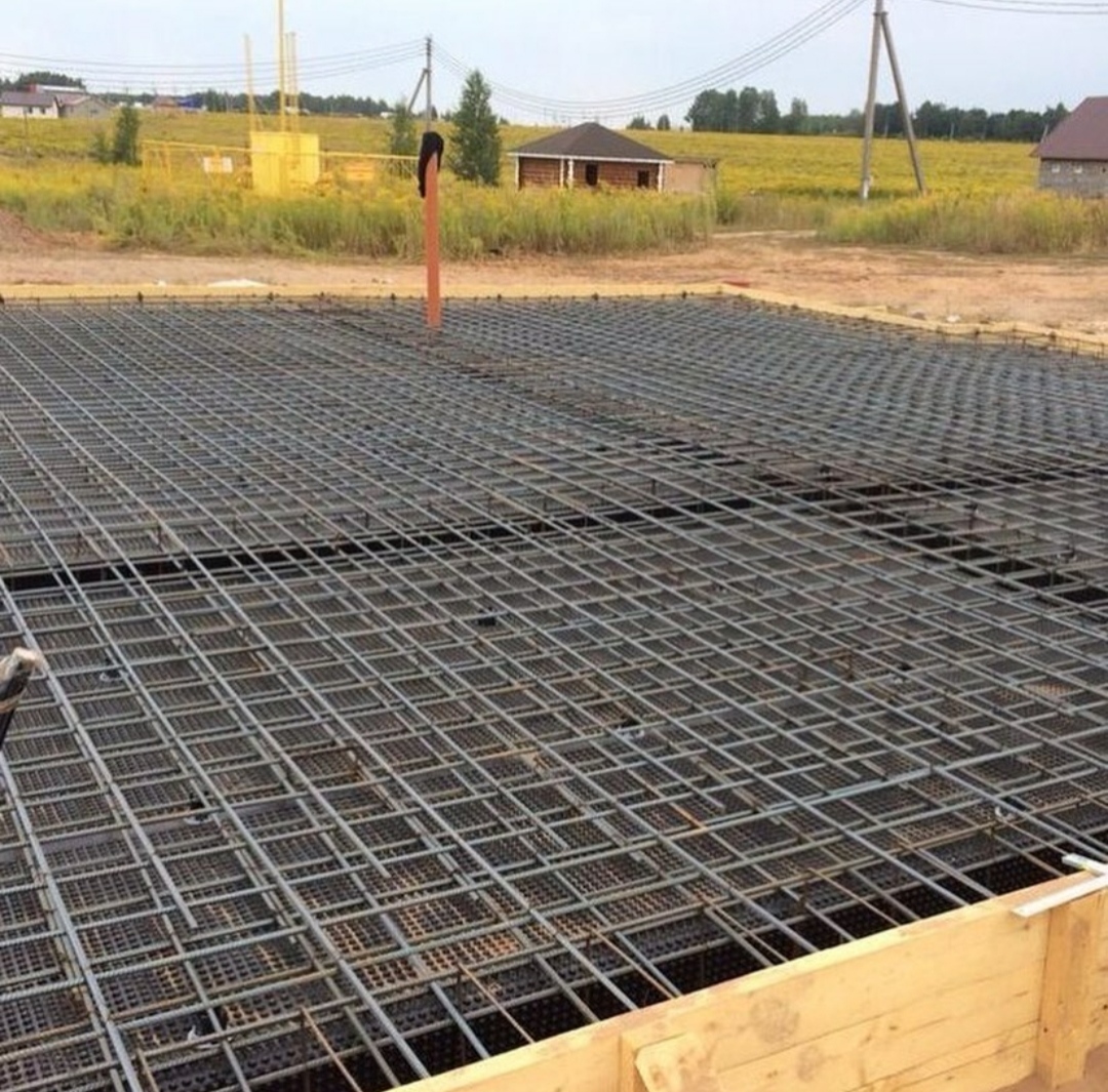 Zbrojenie fundamentu domu jednorodzinnego z widoczną siatką stalową i szalunkiem drewnianym, na tle krajobrazu z polami i zabudowaniami.