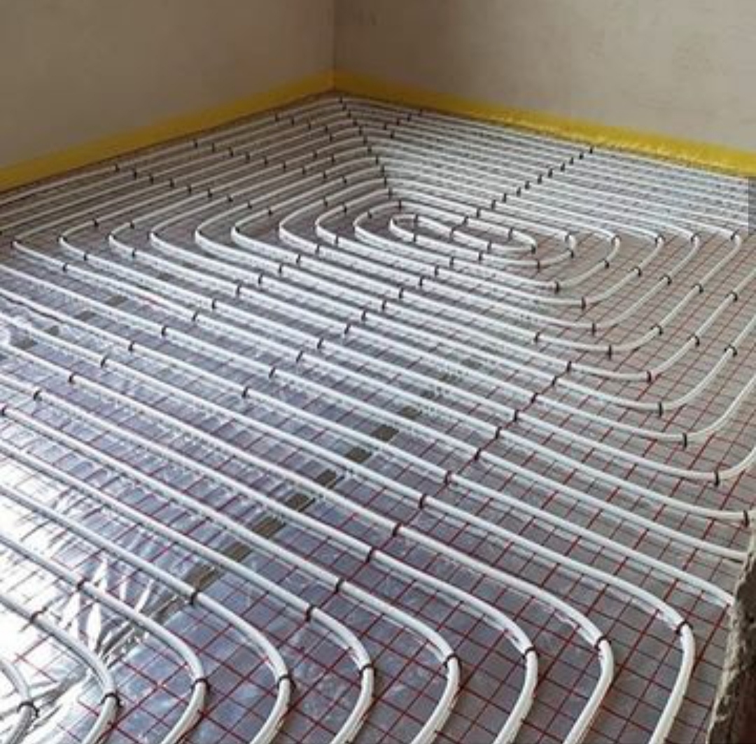 Instalacja ogrzewania podłogowego: białe rurki ułożone w spiralny wzór na srebrnej folii z czerwoną siatką, widoczna żółta taśma maskująca przy ścianie.