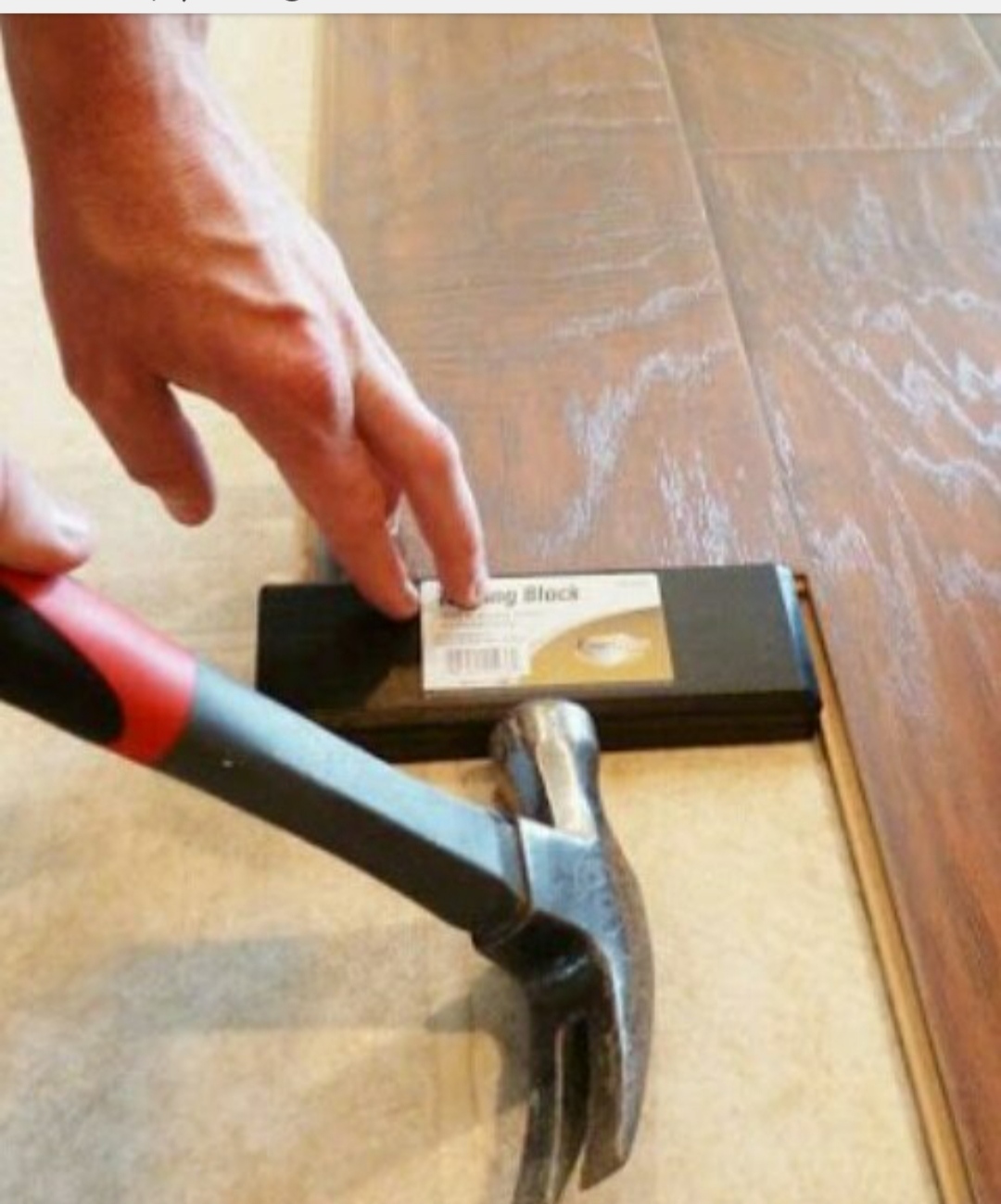 Instalacja paneli podłogowych: dłoń dociska klocek dobijający, a młotek z czerwono-czarną rękojeścią przygotowany do użycia, widoczny fragment ułożonej podłogi.
