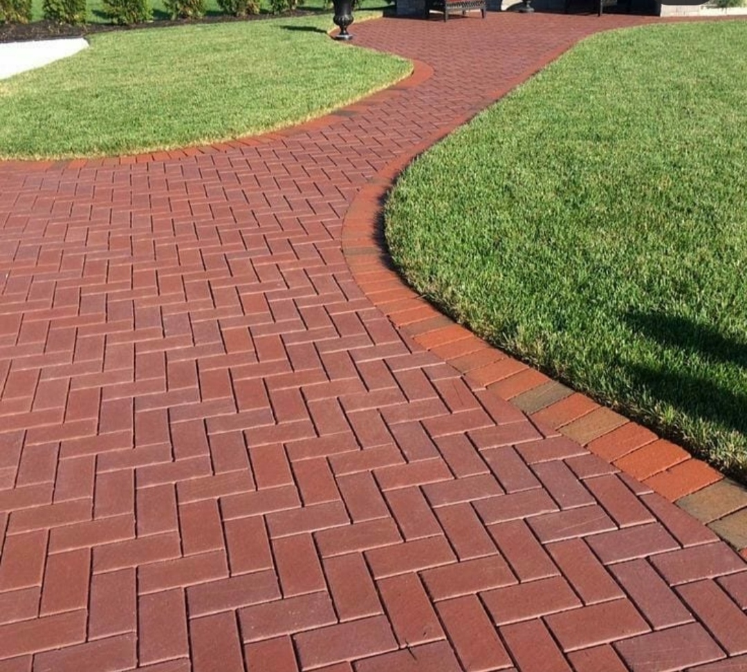 Czerwona ścieżka z cegły ułożona w jodełkę, obrzeża z pojedynczych cegieł, trawnik po bokach, w oddali ławka i donica.