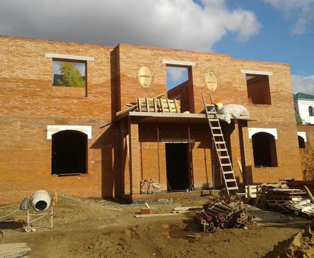 Wznoszenie dwukondygnacyjnego domu z czerwonej cegły, widoczne otwory okienne i drzwiowe, drewniane rusztowanie, drabina oparta o ścianę, betoniarka i materiały budowlane na placu budowy...