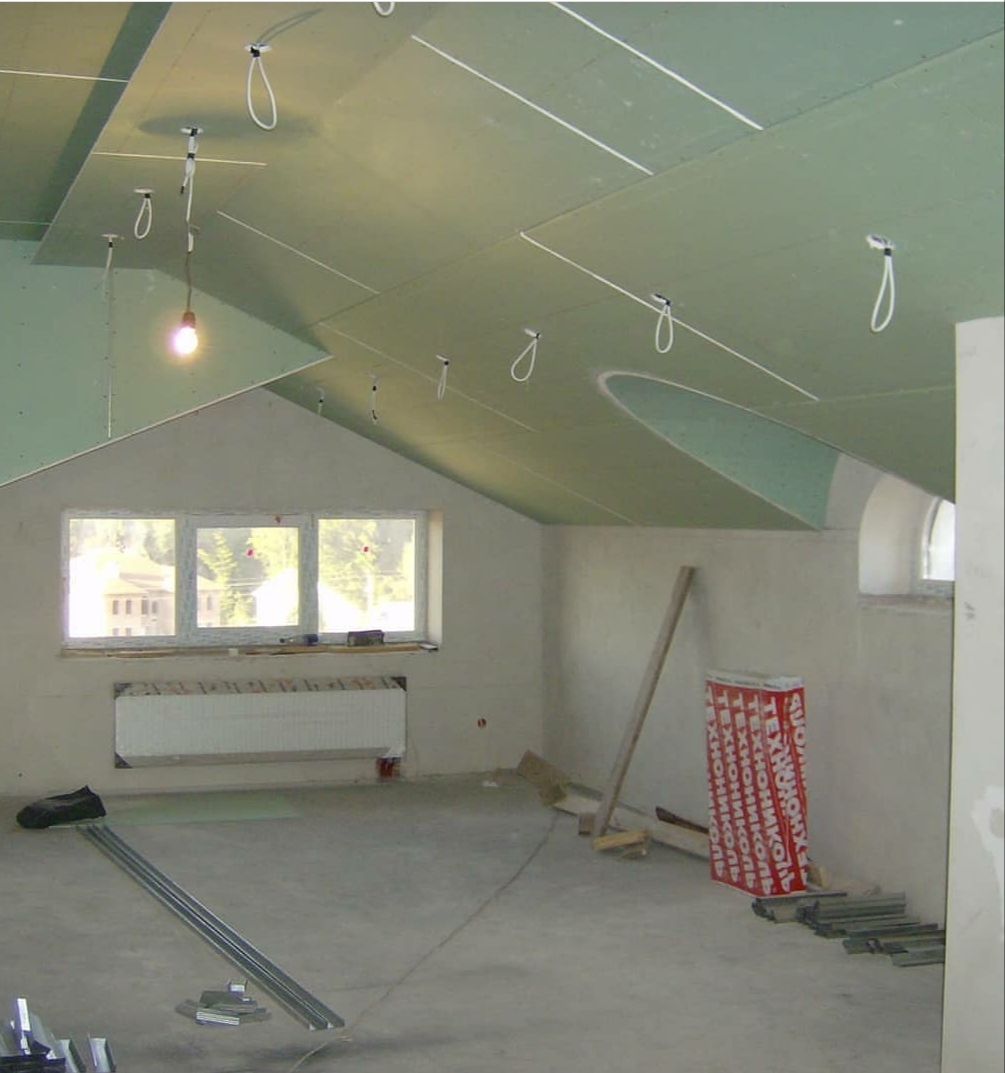 Poddasze w trakcie wykańczania, widoczne zamontowane płyty gipsowo-kartonowe na suficie z przygotowanymi punktami oświetleniowymi, okno dachowe, grzejnik i materiały budowlane na podłodze.