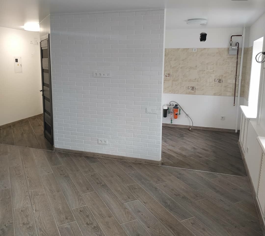 Wnętrze po remoncie: ściana z białej cegły, drewnopodobna podłoga ułożona w jodełkę, widok na kuchnię z beżowymi płytkami i filtrami do wody.