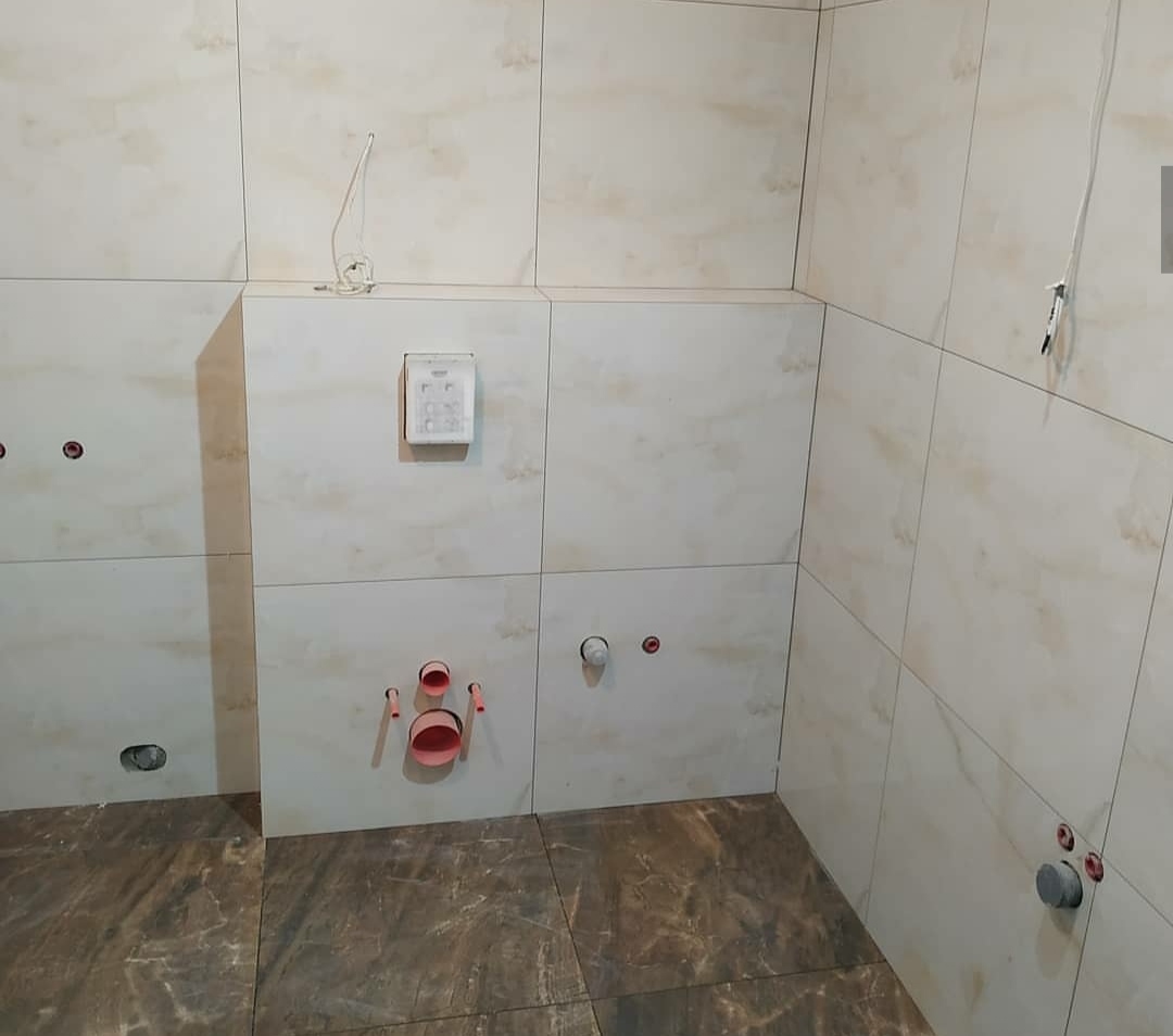 Wnętrze łazienki w trakcie remontu, widoczne kremowe płytki na ścianach, brązowe płytki na podłodze, wystające rury i kable, instalacja podtynkowego WC w trakcie montażu.