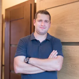 Uśmiechnięty mężczyzna w granatowej koszulce polo, z zegarkiem na ręku, stoi na tle dwóch różnych modeli drzwi wewnętrznych.