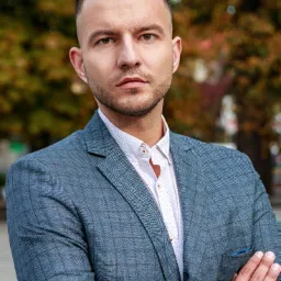 Portret mężczyzny w niebieskiej marynarce w kratę i białej koszuli, z rękami skrzyżowanymi na piersiach, na tle rozmytych drzew w jesiennych barwach; fotografia wykonana we Wrocławiu.