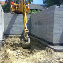 Żółta koparka na placu budowy, przygotowująca teren pod fundamenty, widoczne betonowe ściany szalunkowe i wykopany grunt.