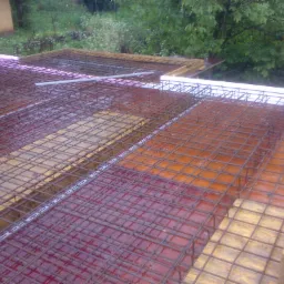 Zbrojenie stropu w trakcie budowy, widoczne drewniane szalunki, siatka zbrojeniowa, styropianowe obrzeża i elementy instalacji, tło z zielenią.