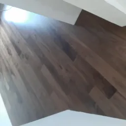 Układanie paneli podłogowych w odcieniach brązu, widoczne białe listwy przypodłogowe oraz fragment okna i ściany w trakcie prac wykończeniowych.