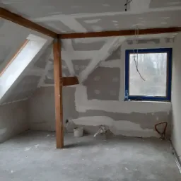 Poddasze w trakcie prac wykończeniowych: widoczne płyty gipsowo-kartonowe na skosach dachu, drewniana konstrukcja wspierająca, okno zabezpieczone taśmą malarską, wystające przewody elektryczne...