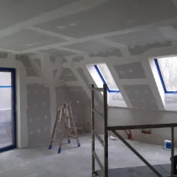 Poddasze w trakcie szpachlowania i przygotowywania pod malowanie, widoczne płyty kartonowo-gipsowe na skośnym suficie i ścianach, okna dachowe zabezpieczone folią malarską, drabina i rusztowanie...