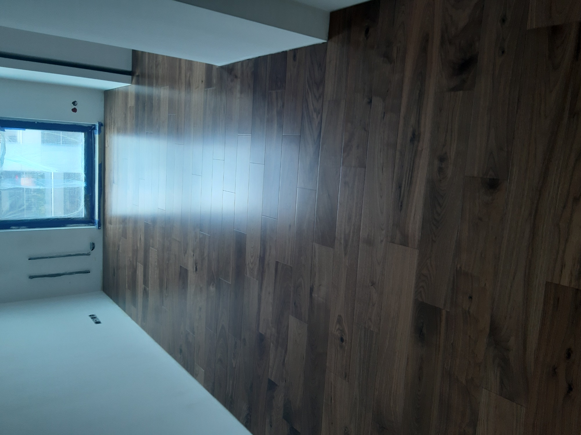 Świeżo położona, ciemna okładzina ścienna imitująca drewno w surowym pomieszczeniu z oknem zabezpieczonym folią, widoczne wystające przewody elektryczne.