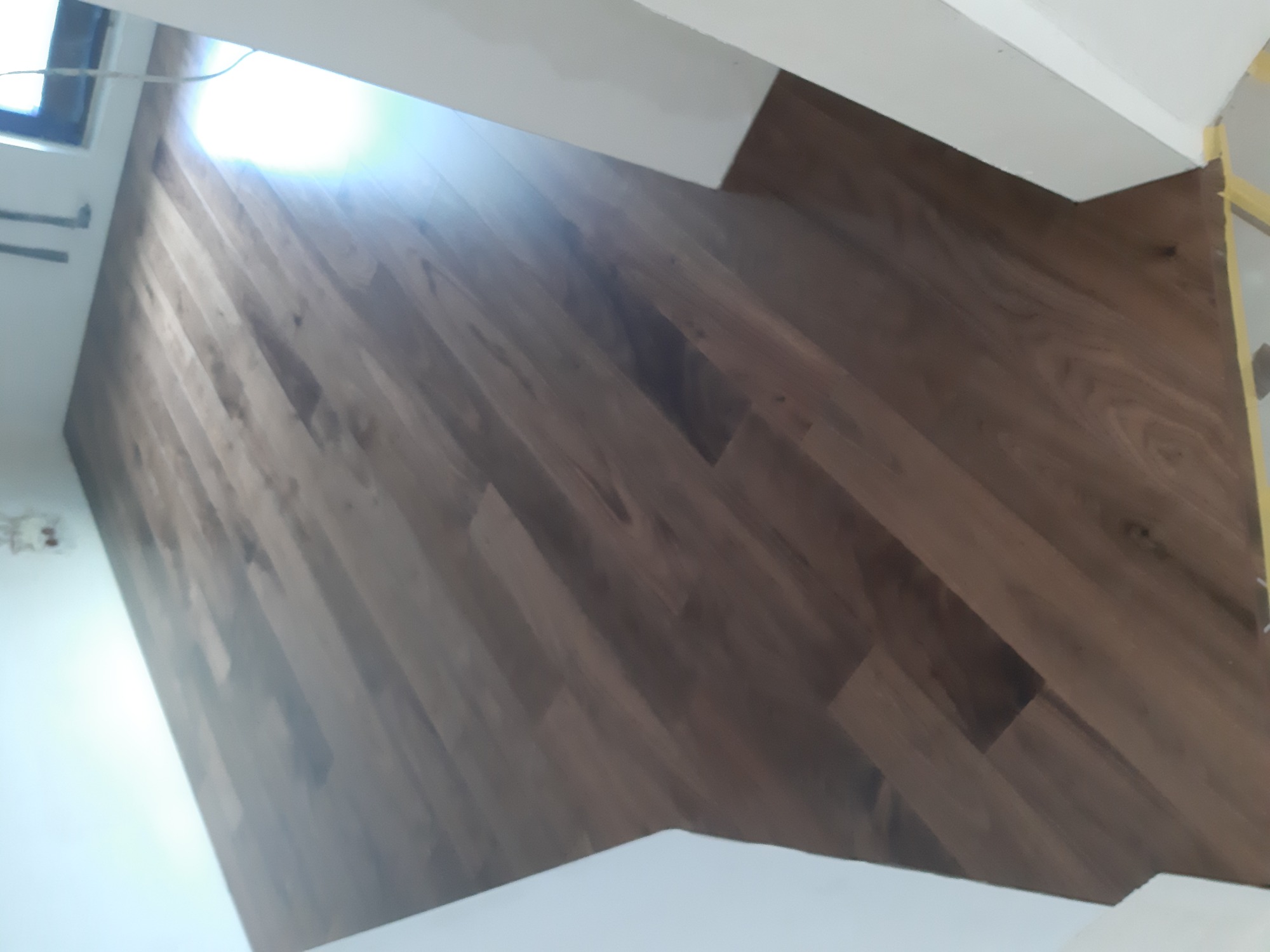 Układanie paneli podłogowych w odcieniach brązu, widoczne białe listwy przypodłogowe oraz fragment okna i ściany w trakcie prac wykończeniowych.