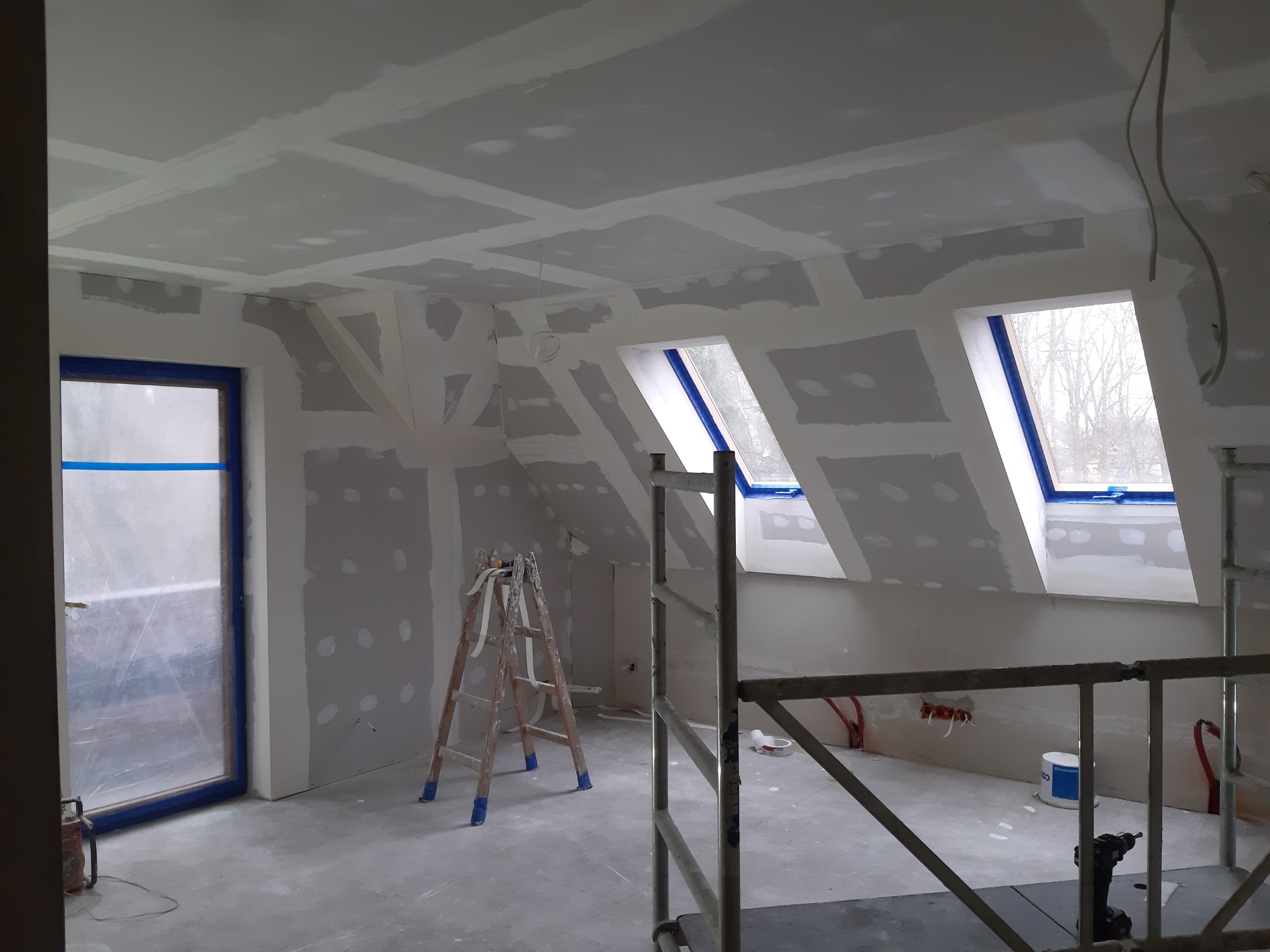 Poddasze w trakcie szpachlowania i przygotowywania pod malowanie, widoczne płyty kartonowo-gipsowe na skośnym suficie i ścianach, okna dachowe zabezpieczone folią malarską, drabina i rusztowanie...