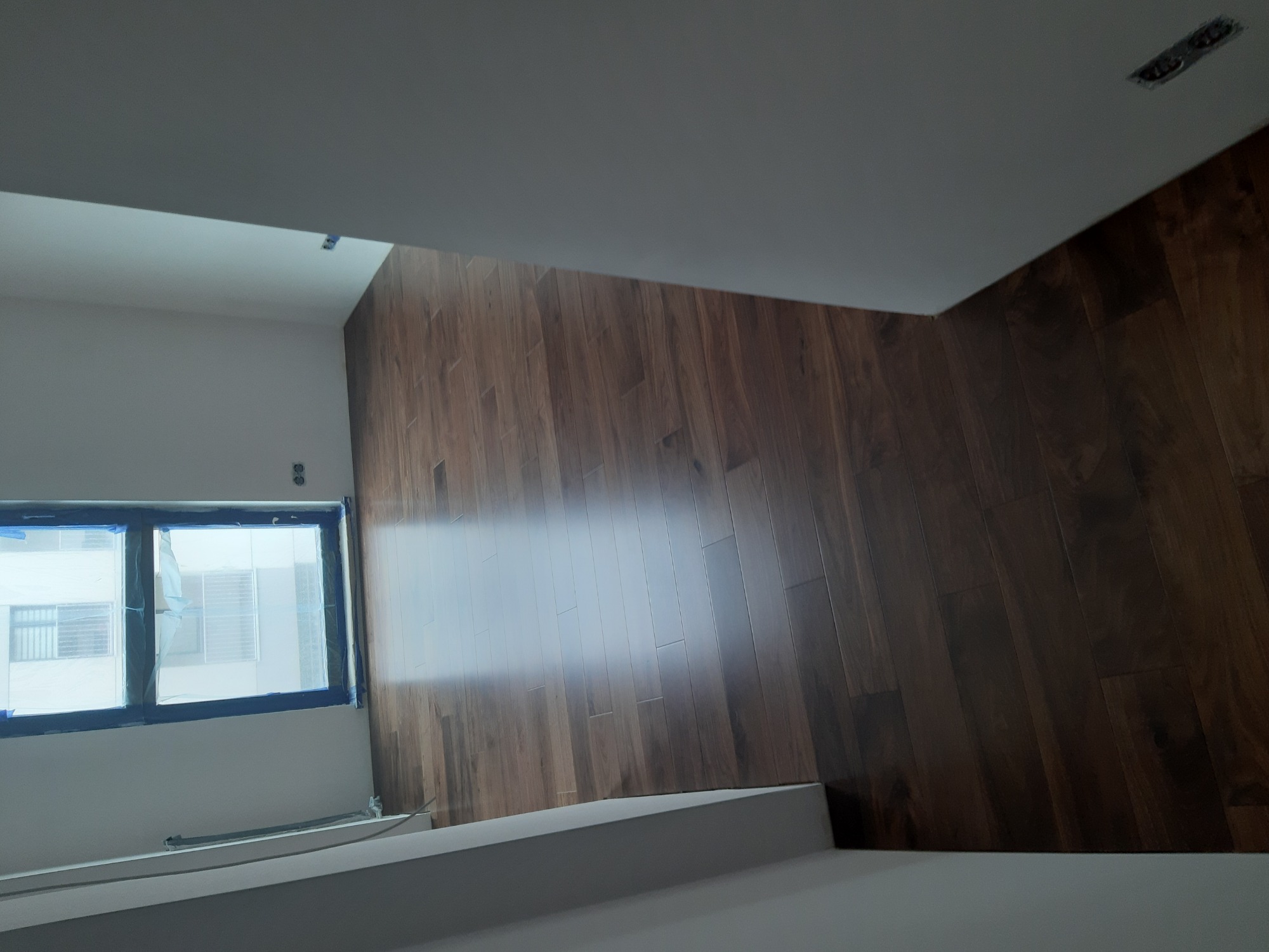 Ściana wykończona panelami drewnopodobnymi, okno zabezpieczone folią malarską, widoczne przygotowania do prac wykończeniowych.