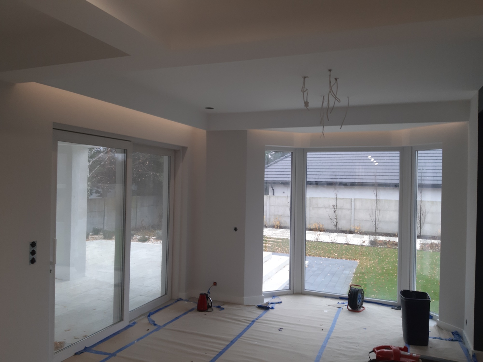 Jasne wnętrze w trakcie prac przygotowawczych: podłoga zabezpieczona folią i taśmą malarską, widoczne okna z widokiem na ogród, wystające kable oświetleniowe z sufitu, ściany przygotowane...