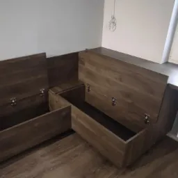 Drewniana zabudowa z pojemnikami z otwieranymi wiekami, zamontowana przy oknie, widoczne metalowe zatrzaski na wiekach.