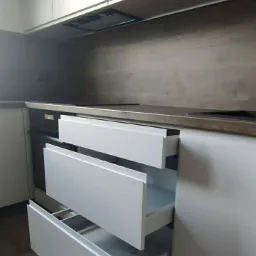 Nowoczesna kuchnia z białymi, bezuchwytowymi frontami szuflad, z których trzy są wysunięte, ukazując półki wewnątrz. Blat w kolorze ciemnego drewna kontrastuje z jasnymi szafkami.