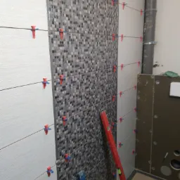 Układanie płytek mozaikowych i białych płytek z poziomnicą i systemem poziomowania płytek w trakcie remontu łazienki.