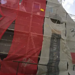 Dom w trakcie prac elewacyjnych, osłonięty rusztowaniami z czerwoną i białą siatką ochronną, widoczny fragment ściany z szarymi płytami izolacyjnymi.