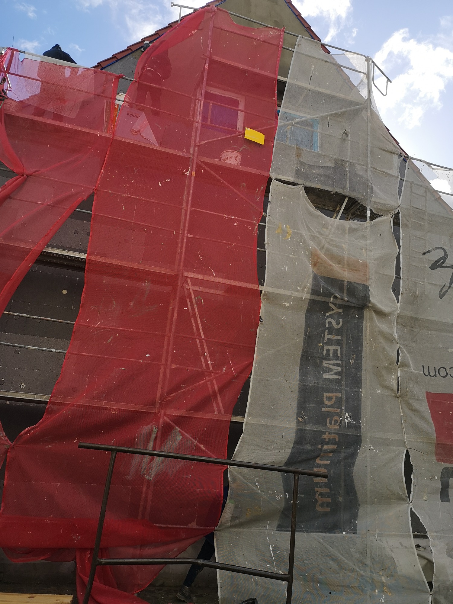 Dom w trakcie prac elewacyjnych, osłonięty rusztowaniami z czerwoną i białą siatką ochronną, widoczny fragment ściany z szarymi płytami izolacyjnymi.