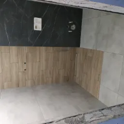 Wykończona łazienka z czarną marmurową ścianą, drewnopodobnymi płytkami na ścianie obok i szarymi płytkami na podłodze i ścianie, widoczne otwory na armaturę i odpływ liniowy.