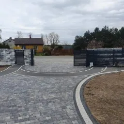 Ułożony podjazd z szarej kostki brukowej z obramowaniem z jasnych kostek, prowadzący do automatycznej bramy wjazdowej, widok na dom w tle pod pochmurnym niebem.