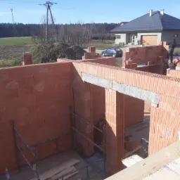 Wznoszenie ścian parterowego domu z czerwonej ceramiki poryzowanej, widoczne zbrojenie nad otworem okiennym, w tle drugi budynek z szarą dachówką i błękitny samochód osobowy.