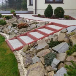 Ogród z kamiennymi schodami i ścieżką z jasnego kruszywa. Widoczne krzewy i trawnik. Dom w tle. Dekoracyjne kamienie tworzą skalniak.