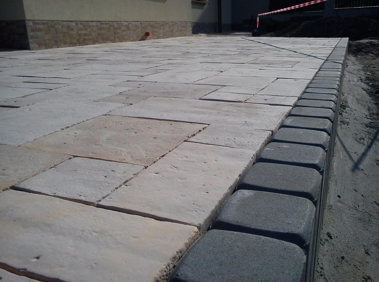 Jasna kostka brukowa ułożona na posesji, obrzeża z ciemnej kostki. Widok z góry, w tle budynek. Kostka o nieregularnym kształcie, tworzy ciekawą fakturę.