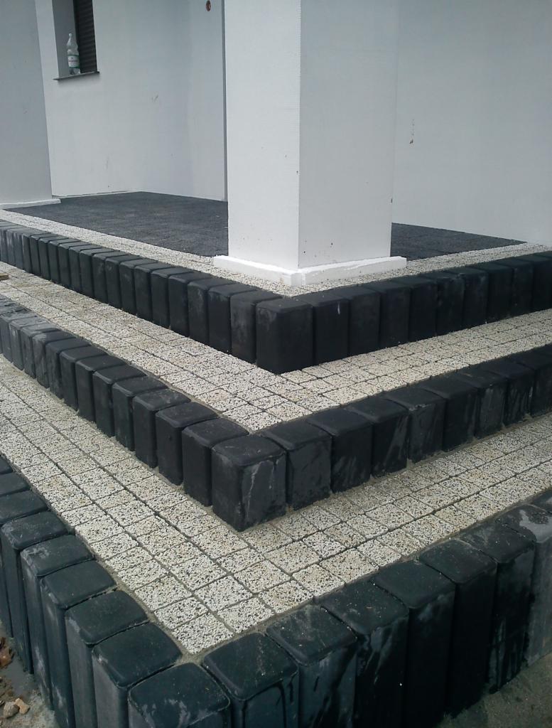 Schody wejściowe wyłożone kostką brukową w odcieniach szarości, obrzeża z ciemnej kostki, widok z dołu, z kolumną budynku w tle.