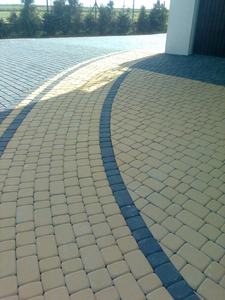 Żółta kostka brukowa ułożona łukiem, obrzeża z ciemnej kostki, w tle brama garażowa i zieleń. Widoczne delikatne cieniowanie.