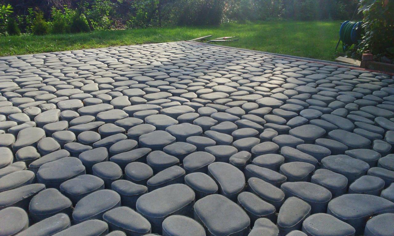 Szara nawierzchnia z kostki brukowej imitującej kamienie rzeczne, otoczona zielenią trawnika. Ujęcie z niskiej perspektywy podkreśla fakturę i regularność ułożenia.