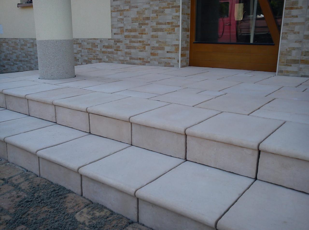 Jasne, równe stopnie z kostki brukowej prowadzące do drzwi wejściowych, z kolumną po lewej stronie. Widoczny fragment muru z cegły na ścianie budynku.