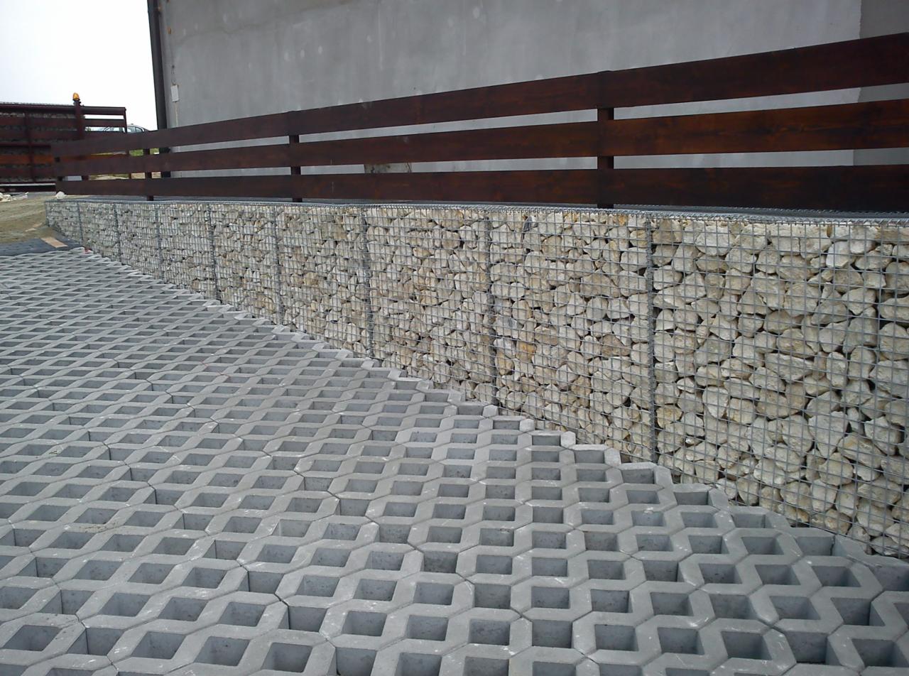 Szara ażurowa kostka brukowa na podjeździe, obok mur z kamieni w metalowej siatce i drewniane ogrodzenie. Widok z góry.