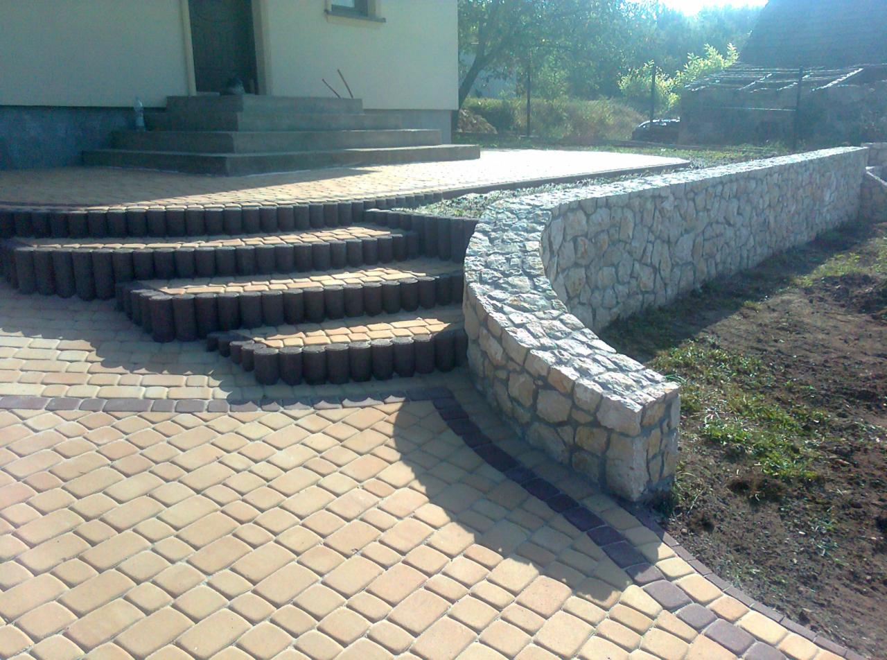 Ułożona kostka brukowa w odcieniach beżu i brązu tworzy ścieżkę i schody. Kamienny murek w tle. Całość tworzy estetyczny krajobraz wokół domu.