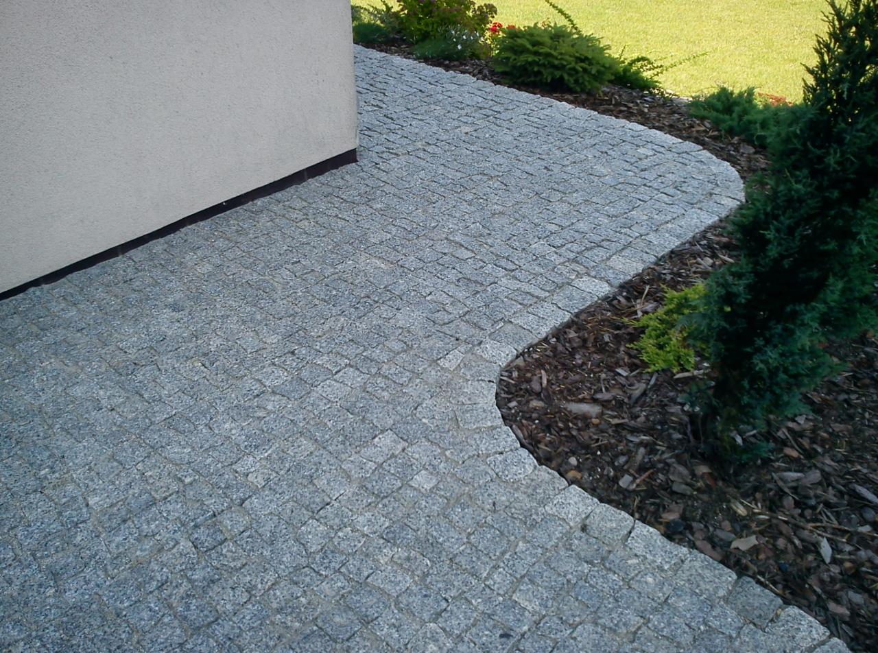 Szara kostka granitowa ułożona wzdłuż ściany budynku i rabaty z korą, tworząca ścieżkę ogrodową z roślinnością iglastą w tle.