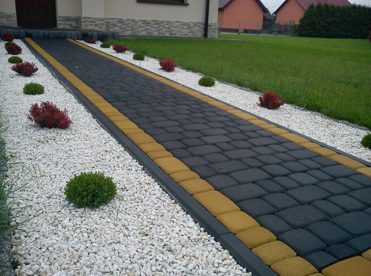 Wyłożony kostką brukową chodnik z obrzeżami w kolorze piaskowym, otoczony białym grysem i niskimi krzewami, prowadzący do domu z trawnikiem w tle.