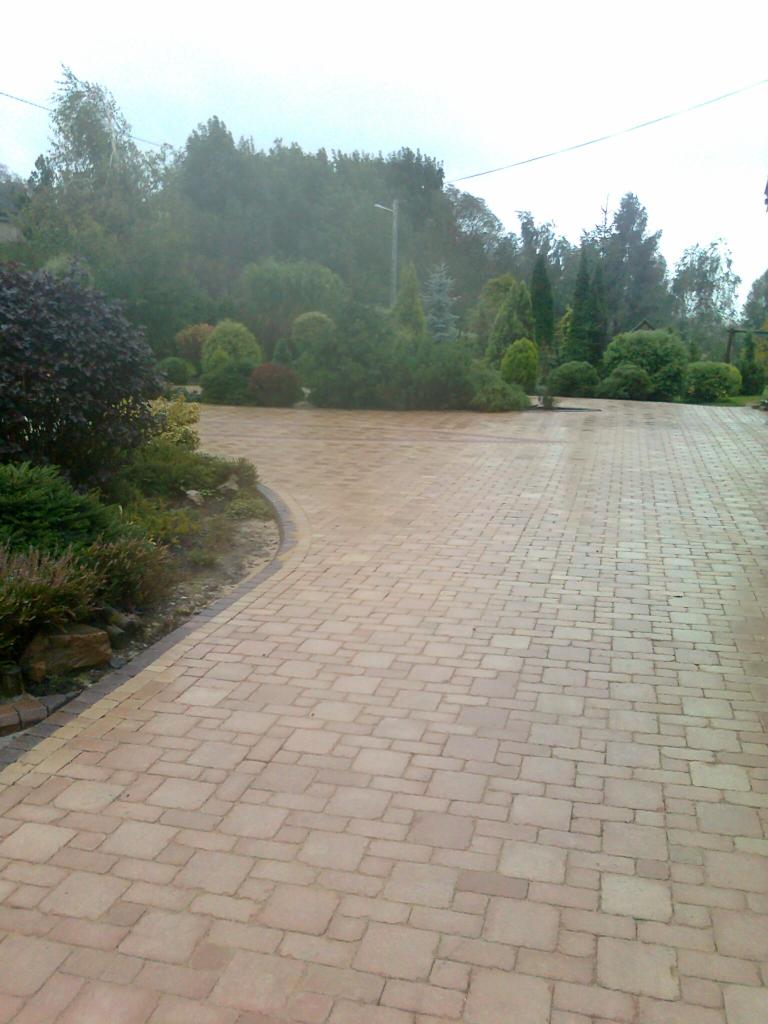 Kaskadowy układ beżowej kostki brukowej tworzy ścieżkę przez zadbany ogród z krzewami i drzewami. Widok na łagodny spadek terenu.