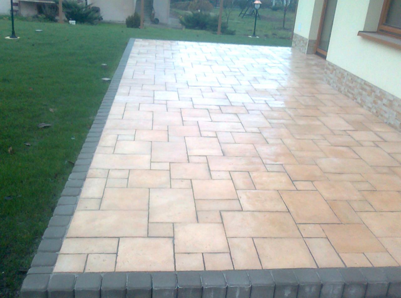 Jasna kostka brukowa ułożona na tarasie przy domu, obrzeża z szarej kostki. Widok na trawnik i elementy ogrodu.