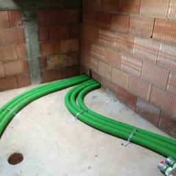 M. WENT Marek Kawka - Zielone karbowane rury wentylacyjne prowadzone wzdłuż surowej ceglanej ściany wewnątrz budynku w stanie deweloperskim, zabezpieczone metalowymi opaskami.