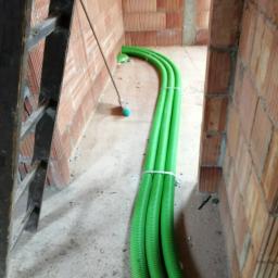 M. WENT Marek Kawka - Trzy zielone rury wentylacyjne, spięte opaskami, ułożone w surowym wnętrzu budynku z cegły, obok drabiny i miotły.