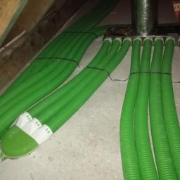 M. WENT Marek Kawka - Zielone, karbowane rury wentylacyjne podłączone do metalowej rozdzielnicy w przestrzeni poddasza, zabezpieczone opaskami. Widoczne drewniane elementy konstrukcji dachu.