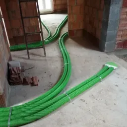 Wnętrze budynku w stanie surowym z widoczną instalacją rekuperacji: zielone, karbowane rury prowadzone po podłodze, mocowane metalowymi opaskami, z widoczną drabiną opartą o ścianę z cegły.