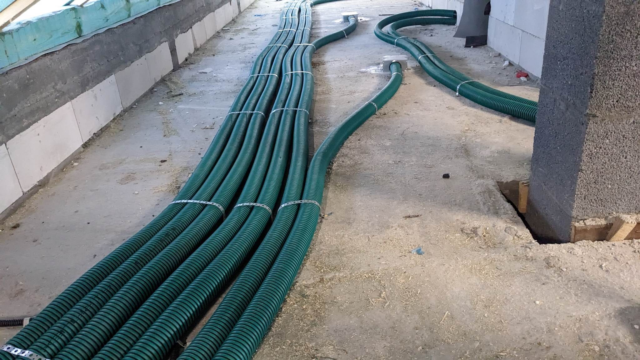 Zielone, karbowane rury wentylacyjne ułożone równolegle na surowym betonowym podłożu w trakcie montażu, zabezpieczone metalowymi opaskami.