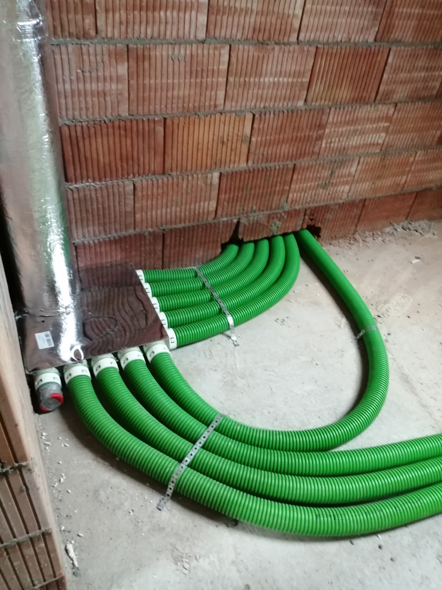 Wewnętrzna instalacja wentylacyjna z zielonymi rurami karbowanymi w surowym stanie, widoczne połączenia z centralą i przejście przez ścianę z cegły dziurawki.