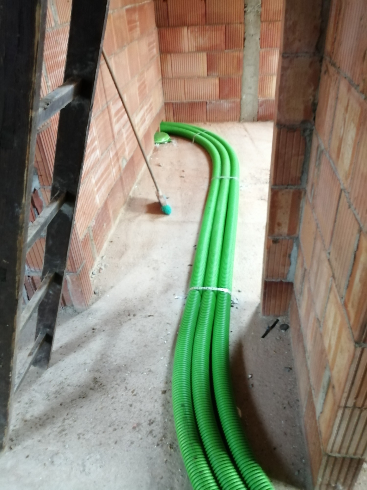 Trzy zielone rury wentylacyjne, spięte opaskami, ułożone w surowym wnętrzu budynku z cegły, obok drabiny i miotły.