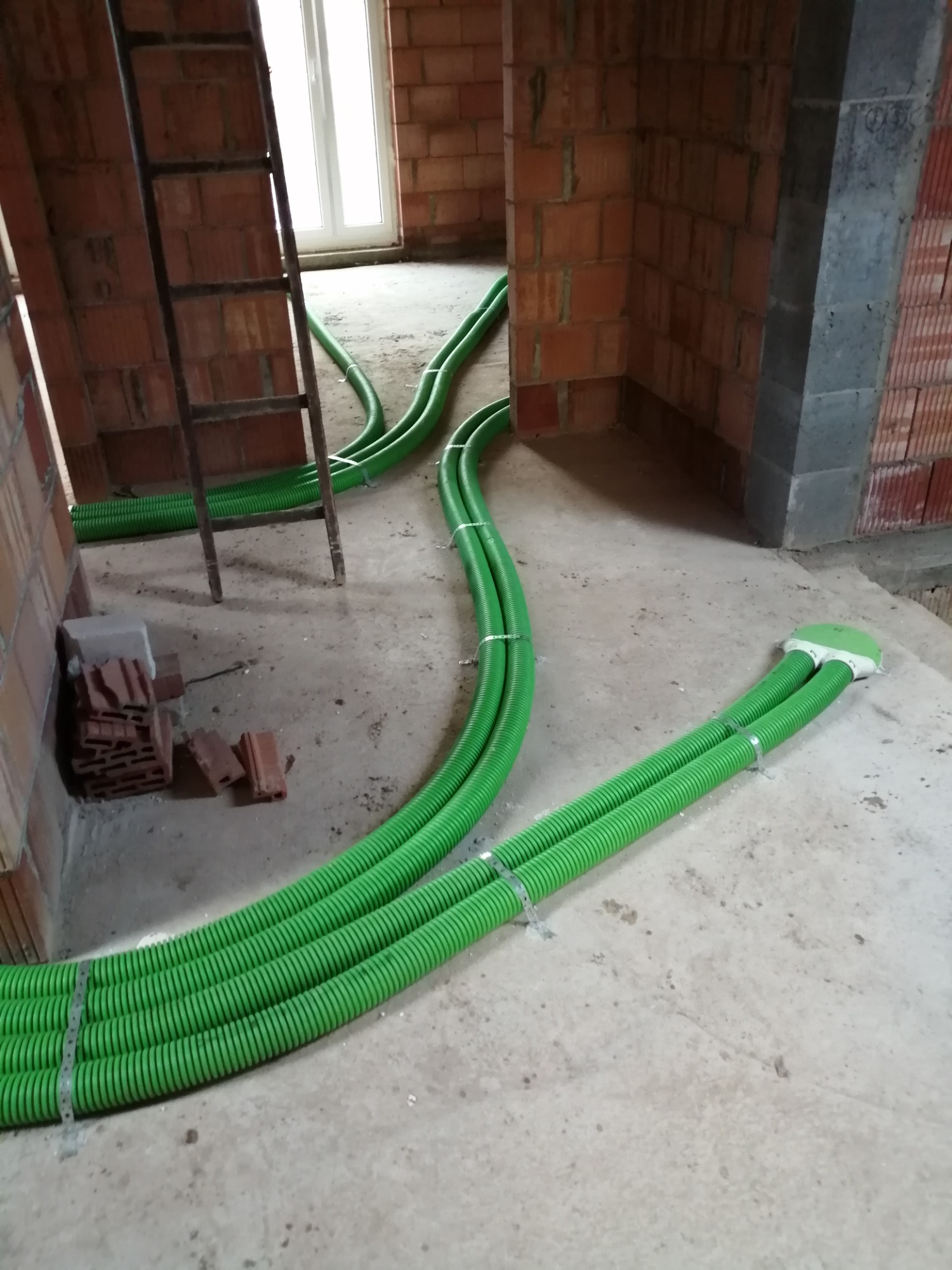 Wnętrze budynku w stanie surowym z widoczną instalacją rekuperacji: zielone, karbowane rury prowadzone po podłodze, mocowane metalowymi opaskami, z widoczną drabiną opartą o ścianę z cegły.
