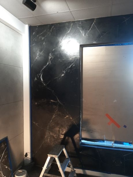 Ściana wykończona czarnym tynkiem imitującym marmur, zabezpieczone okno folią malarską, drabina aluminiowa w pomieszczeniu z szarą ścianą po lewej.
